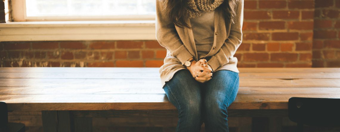 A person sitting quietly on a wooden bench by a window with hands clasped