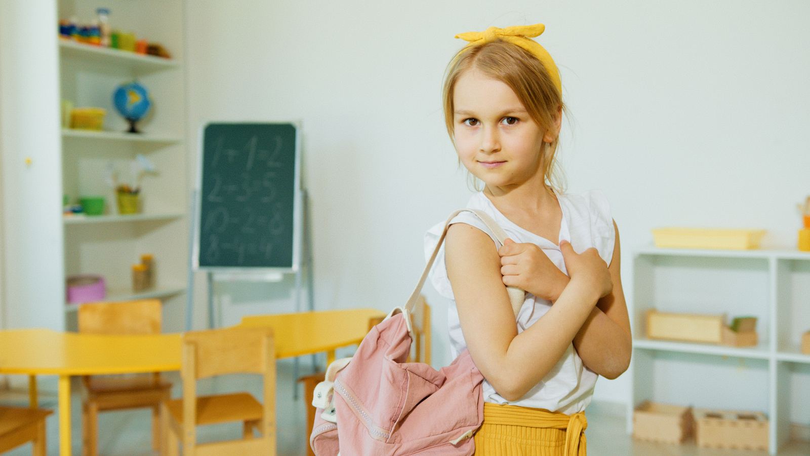 A young child standing in a bright classroom holding a backpack