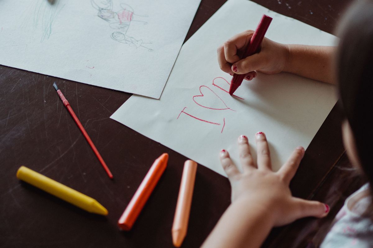 A child's hand holding a crayon beside coloured pencils and paper