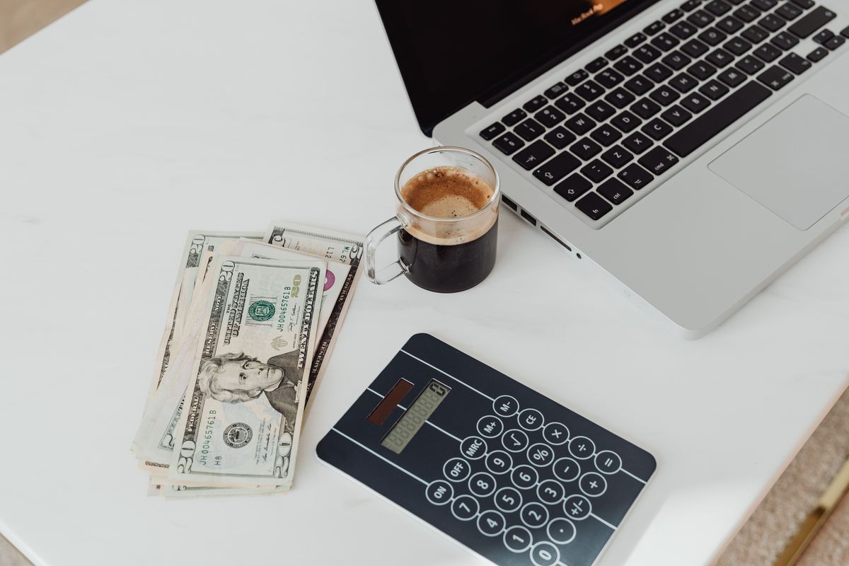 A desk with a calculator, cash, a laptop and a cup of coffee