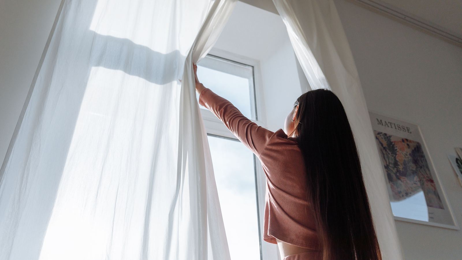 A person opening white curtains to let daylight into a room