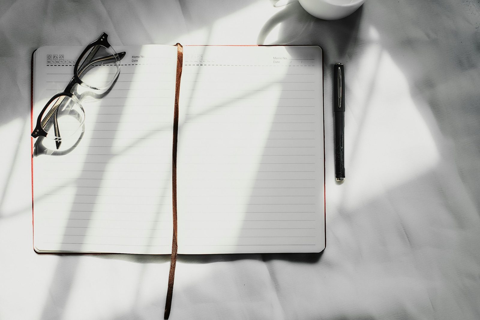 An open notebook with a pen and reading glasses on a desk in bright natural light