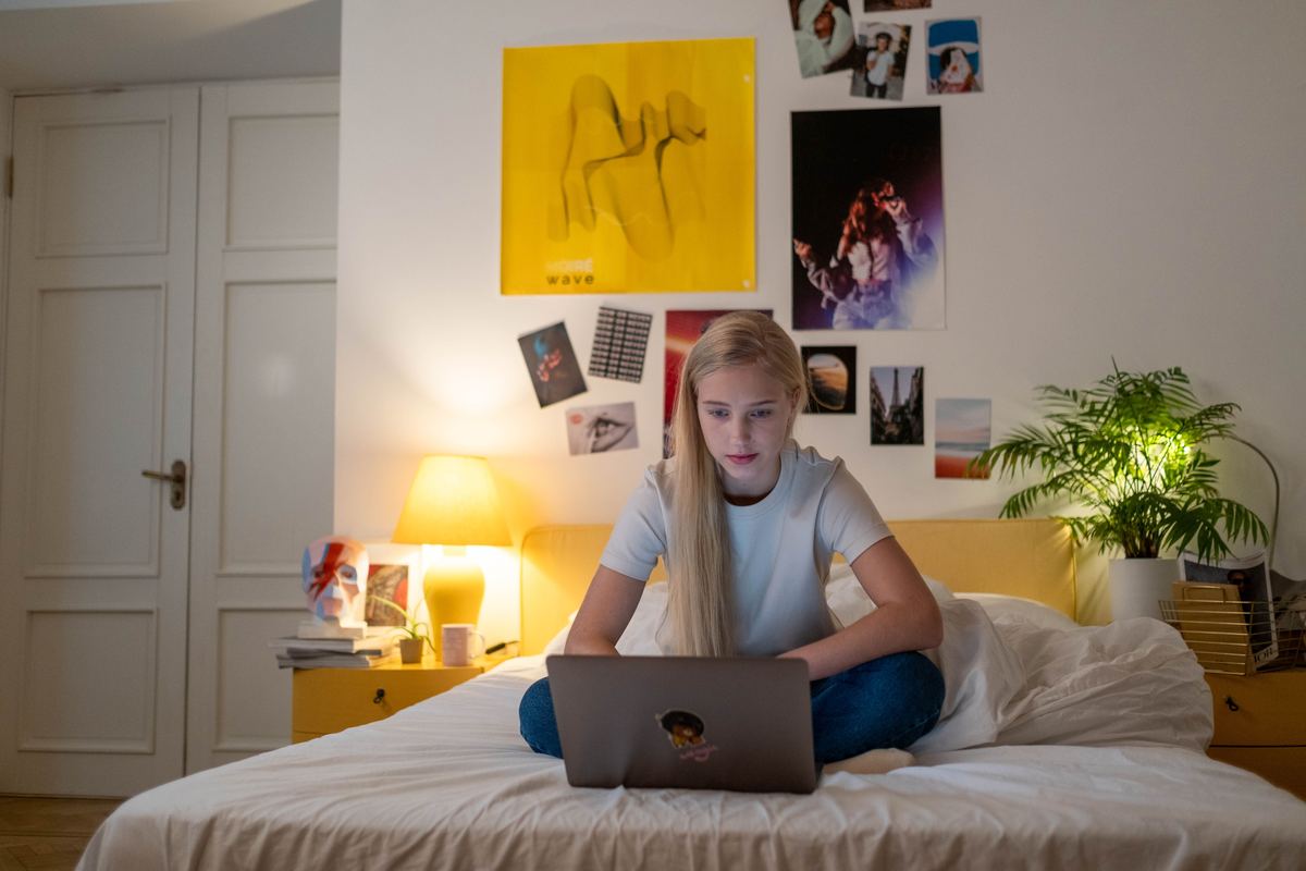 A teenager using a laptop on a bed in a warmly lit room