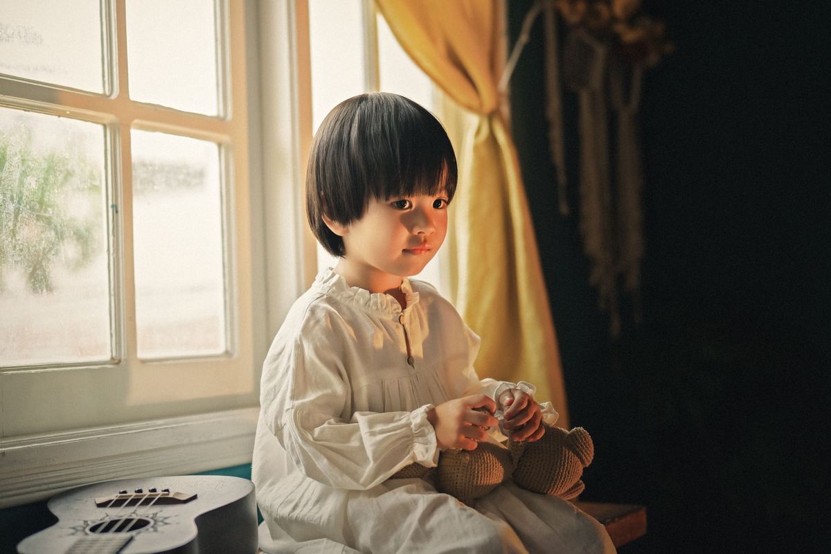 A child sitting by a window holding a soft toy