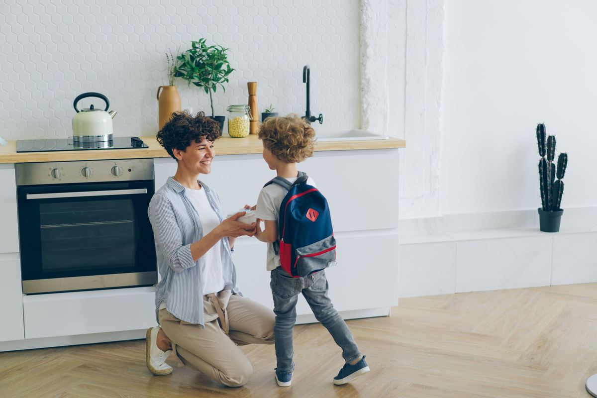 A parent crouching to help a child with a backpack in a kitchen