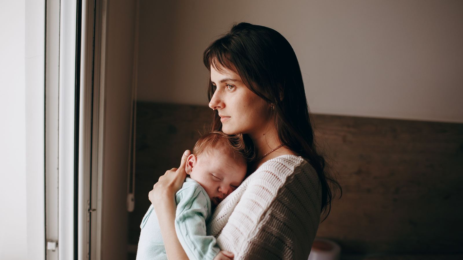 A parent holding a sleeping baby beside a window